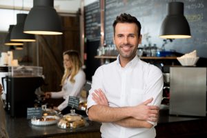 Business owner smiling and standing with arms crossed with an employee working in the background.